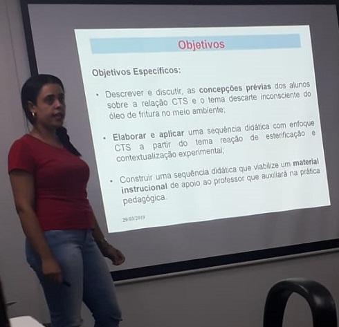 Aluna Juliana Fernandes Lima (morena, blusa vermelha, calça jeans, cabelo castanho preso, de pé, próxima ao slide de apresentação